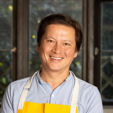 Photo of Kenji Lopez Alt, a man with dark hair wearing a light blue shirt and a yellow apron with white straps.